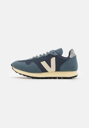 Blå og marineblå sneaker med cremefarvede snørebånd, et stort cremefarvet "V" logo på siden og en lys beige sål på en hvid baggrund.