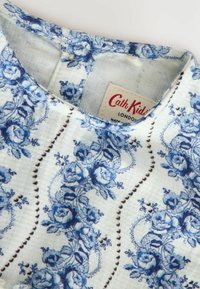 Šviesiai mėlynas audinys su tamsiai mėlynomis gėlių raštais, turintis suapvalintą iškirptę ir mažą etiketę, ant kurios užrašyta "Cath Kidston, London."