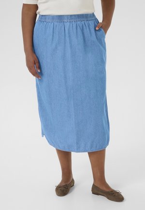 Person iført et knælængde blåt denimskørt med elastisk talje og brune flade sko, stående med den ene hånd i lommen.