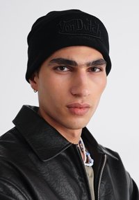 Gorro negro con el logo embosado de "Von Dutch", llevado con una chaqueta de cuero negra. Diseño minimalista, textura suave, forma ajustada.