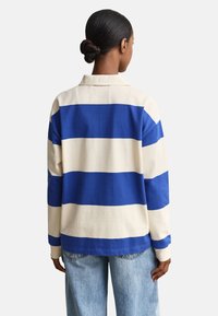 Femme aux cheveux foncés en chignon portant une chemise à manches longues blanche et bleue à rayures horizontales et un jean bleu clair, vue de dos.