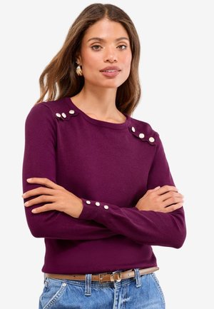 Žena nosící fialový svetr s dlouhým rukávem s dekorativními perlovými knoflíky na ramenech a manžetách, ve spojení s modrými džínami a hnědým páskem.