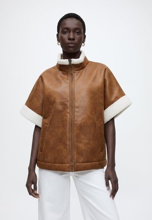 Femme portant une veste en shearling marron à manches courtes avec une doublure blanche, associée à un pantalon blanc, debout devant un fond uni.