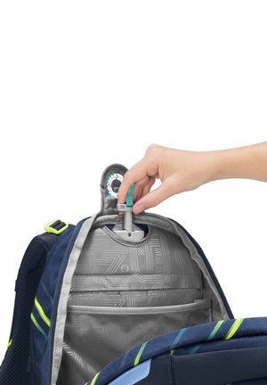 Hand legt einen Hydrationsschlauch in einen dunkelblauen und grauen Rucksack mit limonengrünen Akzenten und einem inneren Fach für eine Hydrationsblase.