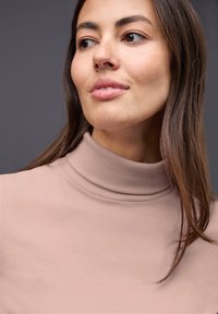 Hoge hals, geribbelde top van zachtroze stof. Heeft een aansluitend ontwerp met een gladde textuur en een close-up van de halslijn en het gezicht.