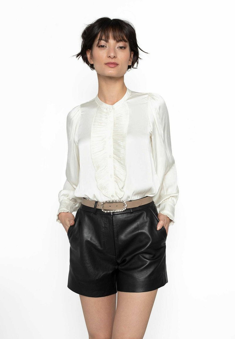 Silkesblus i off-white med vertikala volanger framtill, matchad med svarta lädershorts och ett beige bälte med en dekorativ spänne.