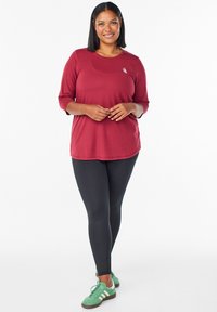 Musta urheiltoppi, jossa on kolme neljännesosaa hihat, yhdistettynä mustiin legginsseihin ja vihreisiin lenkkareihin. Huomattava logodetaili vasemmalla ylhäällä.