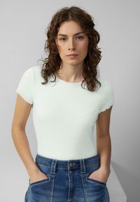 Junge Frau mit lockigem braunem Haar, die ein figurbetontes weißes Kurzarmoberteil und blue Jeans trägt, mit den Händen in den Taschen, vor neutralem Hintergrund.
