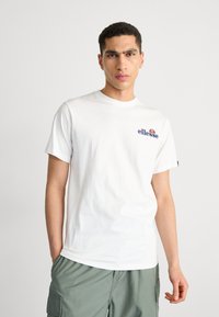 T-shirt bianca in cotone con un piccolo logo "ellesse" blu e arancione sul petto. Maniche corte, colletto girocollo, vestibilità rilassata e senza fantasie.