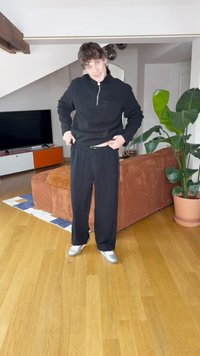 Czarny sweter na suwak z teksturowanym wzorem, zestawiony z luźnymi czarnymi spodniami dresowymi. Stylizację dopełniają połyskujące srebrne buty sportowe.