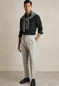 Light gray pleated trousers with side pockets, paired with a black linen button-up shirt and a gray knit sweater draped around the shoulders.