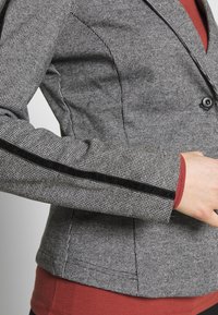 Grå houndstooth blazer med sort velourdetalje på ærmet, har et tætsiddende design, struktureret stof og en enkelt knaplukning.