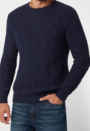 Homme portant un pull en maille torsadée bleu marine avec poignets et ourlet côtelés, associé à un jean bleu, main dans la poche.