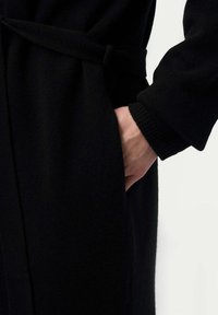 Svart stickad kappa med bälte i midjan, featuring en sidoficka och ribbade ärmar, som betonar den släta texturen och den minimalistiska designen.