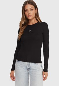 Maglia nera a maniche lunghe con costine e scollo rotondo, con logo bianco "GUESS JEANS" sul petto. Abbinata a jeans chiari.