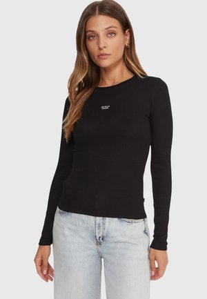 Maglia nera a maniche lunghe con costine e scollo rotondo, con logo bianco "GUESS JEANS" sul petto. Abbinata a jeans chiari.
