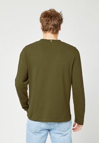 Uomo con capelli castano chiaro che indossa una camicia a maniche lunghe verde oliva e jeans azzurri chiari, di spalle su uno sfondo bianco semplice.
