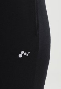 Schwarze Sporthose mit glatter Textur, ausgestattet mit einem kleinen, glänzenden silbernen Logo mit Kreisen in der Nähe der Seitentasche. Einfaches und funktionales Design.