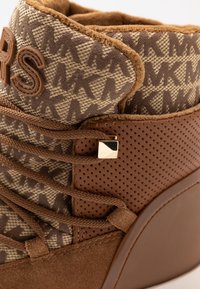 Bottines marron avec un dessus en textile à motifs, des accents en daim, un col rembourré et un hardware doré carré sur les lacets. Détails en cuir perforé.