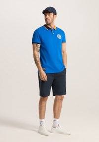 Niebieska koszulka polo z czarnymi i pomarańczowymi detalami, krótkie rękawy i logo. W połączeniu z ciemnymi szortami i białymi trampkami.
