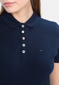 Mörkblå polo skjorta med klassisk krage, sex kontrasterande knappar och en liten logotyp på vänster bröst. Mjuk, texturerad tyg.