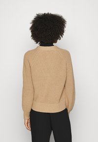 Beige stickad tröja med ett strukturerat mönster, ribbade ärmslut och nederkant, raglanärmar och rund halsringning, bärs över mörka byxor.
