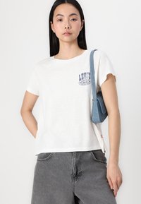 Balts T-krekls ar apaļu kaklu un zilu Levi's logo uz krūts, saskaņots ar pelēkiem denimu šortiem un mazu zilu rokas somiņu.