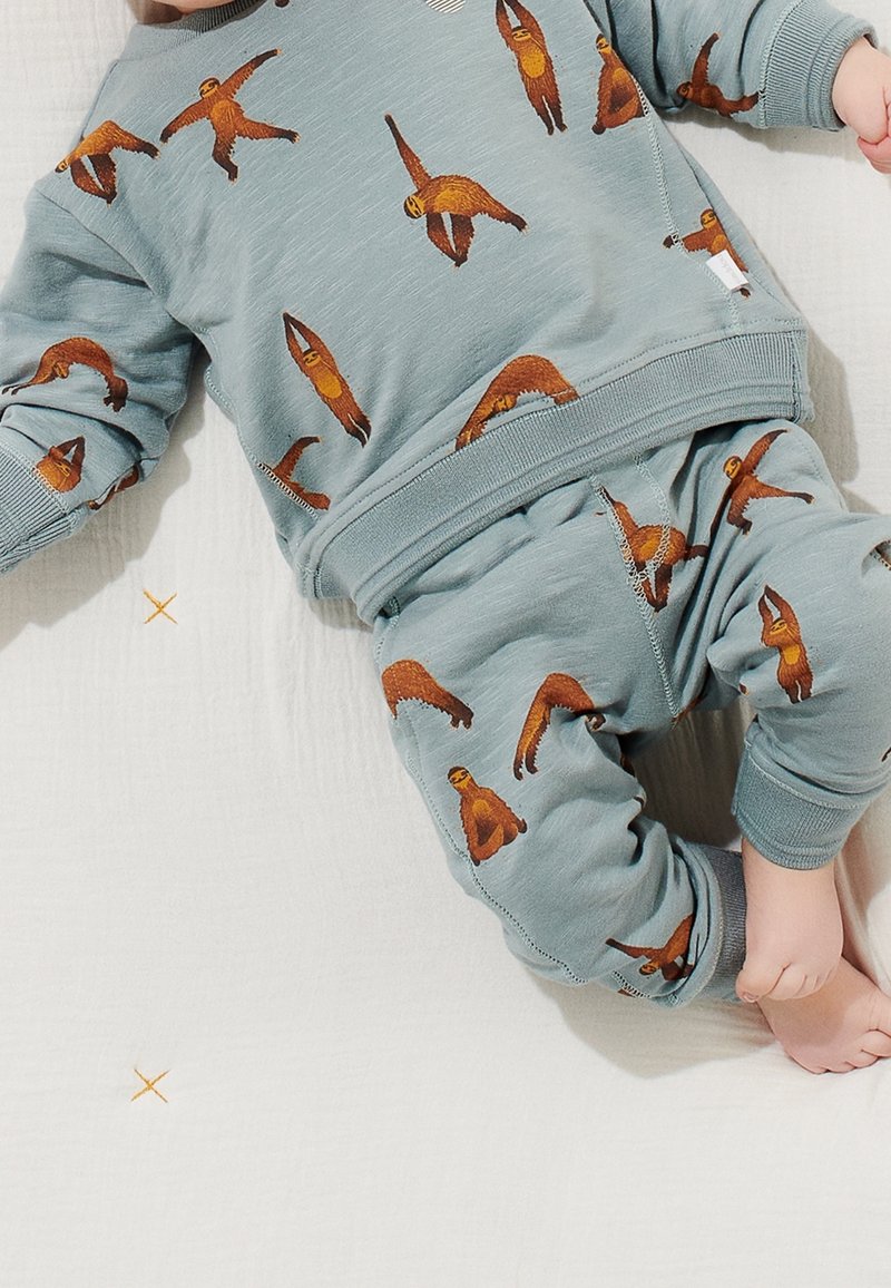 Tout-petit portant une tenue bleu clair avec des motifs ludiques de paresseux orange, allongé sur une surface blanche texturée avec de petites croix brodées.