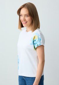 Weißes Baumwoll-T-Shirt mit kurzen Ärmeln, das blaue Blumenmuster auf den Schultern aufweist. Rundhalsausschnitt, lässige Passform und glatte Textur.
