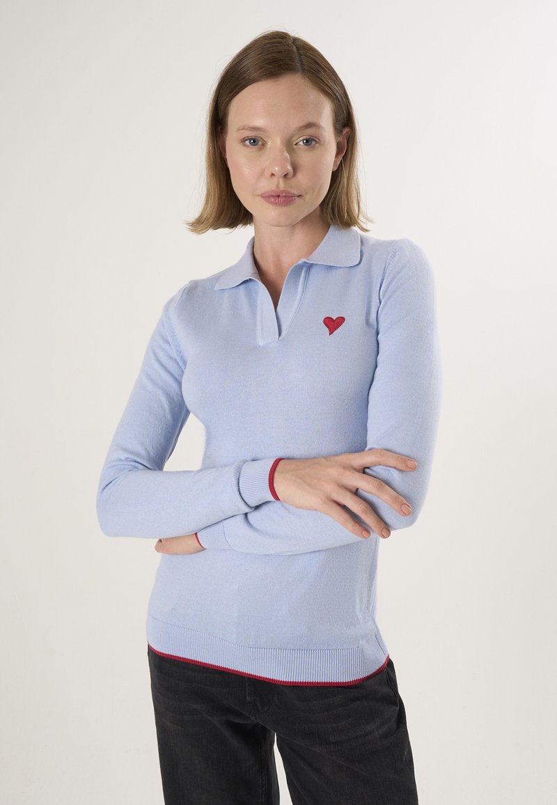 Femme aux cheveux châtain clair portant un haut à manches longues bleu clair avec un emblème de cœur rouge et un pantalon noir, bras croisés sur un fond uni.