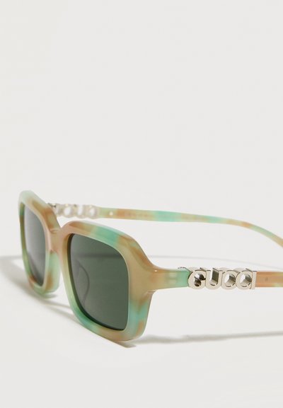 Lunettes de soleil à monture carrée avec un motif marbré vert et beige, verres foncés et logo argenté "Gucci" sur les branches.