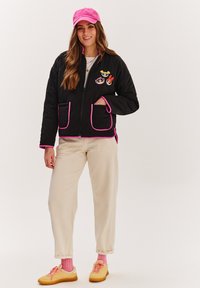 Čierna prešívaná bunda s ružovými akcentmi, zdobená troma vyšívanými kreslenými postavičkami, v kombinácii s béžovými nohavicami, ružovými ponožkami a žltými teniskami.
