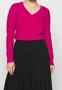 Kobieta ubrana w fuksjowy sweter z dekoltem w serek oraz czarną, wysoką spódnicę plisowaną, z widocznym cienkim złotym naszyjnikiem.