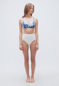 Γυναίκα όρθια, ξυπόλητη, με μπλε φλοράλ μπραλέτα και λευκά εσώρουχα ψηλόμεσα, κοιτάζει προς τα αριστερά της με φόντο απλό.