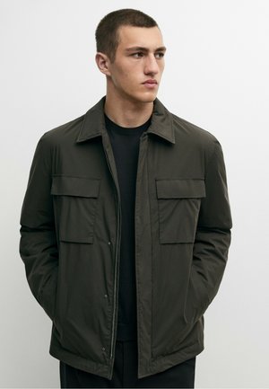 Giovane uomo con capelli corti che indossa una giacca verde scuro con due tasche sul petto, in piedi con le mani nelle tasche, guarda verso destra.
