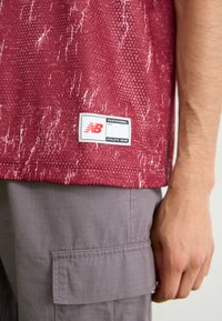 T-shirt de sport rouge avec un design texturé, arborant une étiquette blanche avec le logo "New Balance". Porté avec un pantalon cargo gris.