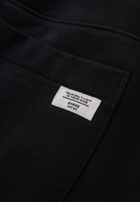 Primer plano de un bolsillo de tela negra con una etiqueta rectangular blanca que dice "TU ÚNICO LÍMITE ES TU MENTE" y "OMBRE desde 1985" cosida.