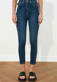 Jeans de mezclilla oscuros de cintura alta con una forma ajustada, que presentan costuras en contraste y una textura suave. Llevados con sandalias deslizantes negras.