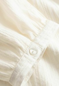 Gros plan d'un poignet en tissu blanc avec un seul bouton blanc et des motifs texturés subtils sur la manche froncée.