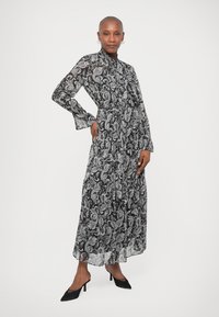 Γυναίκα ποζάρει με ένα μακρύ φόρεμα σε μαύρο και άσπρο με σχέδιο paisley, μακριά μανίκια και μαύρες γόβες με μυτερή μύτη σε άσπρο φόντο.