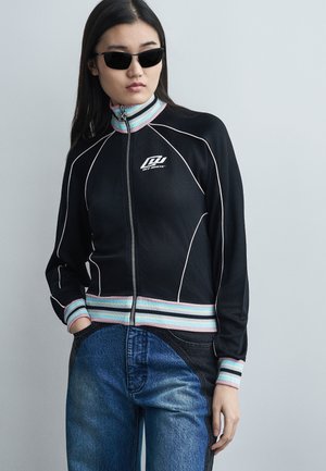 Fiatal nő fekete cipzáras dzsekiben, fehér csíkos díszítéssel, pasztell csíkos gallérral és mandzsettával, kéttónusú kék farmernadrágban, fekete napszemüvegben, kezét a zsebében tartva.