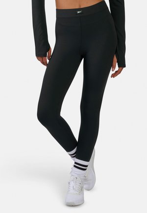 Vrouw draagt zwarte geribbelde legging, witte sneakers en witte sokken met zwarte strepen, staat met gekruiste benen.