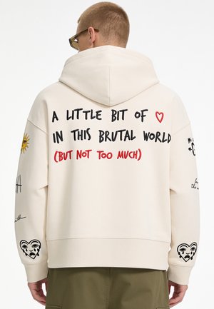 Person trägt einen cremefarbenen Hoodie mit dem Text "A little bit of love in this brutal world (but not too much)" und Herz- sowie Sonnenzeichnungen auf den Ärmeln.