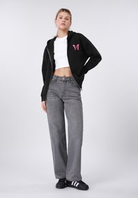 Sweat à capuche noir zippé avec un motif de papillon rose, associé à un jean gris à jambes larges et des baskets noires avec des bandes blanches.