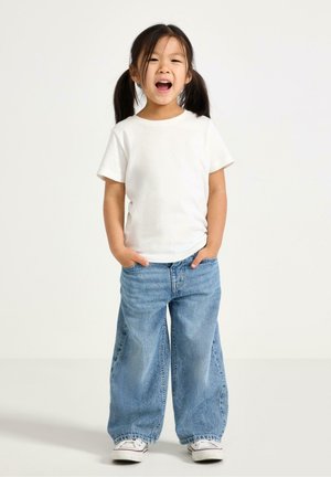 Mladá dívka s tmavými copy stojí s rukama v kapsách, má na sobě bílé tričko, široké modré džíny a bílé tenisky, usmívá se s otevřenými ústy.