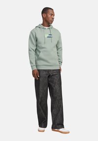 Sweatshirt vert avec une poche avant et un détail de logo, complété par un pantalon en jean foncé. La tenue est accompagnée de baskets blanches avec une semelle marron.