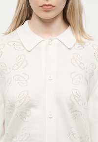 Γυναίκα που φοράει λευκή ζακέτα με κουμπιά, γιακά και διακριτικό λεοπάρ μοτίβο με πλεκτή υφή.