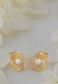 Orecchini a forma di fiore in oro con una perla bianca centrale, caratterizzati da un design texturizzato e dettagli a giorno. Ambientati su uno sfondo morbido.