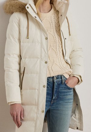 Femme portant un manteau matelassé crème à capuche doublée de fourrure, par-dessus un pull beige en maille torsadée et un jean bleu, une main dans la poche.