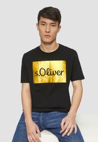 Zwarte t-shirt met een gouden rechthoekig ontwerp met de tekst "s.Oliver" in zwart cursief lettertype. Model gestyled met blauwe jeans.